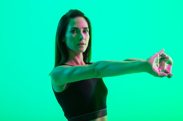 Belle jeune fille de remise en forme debout isolée sur un mur de néon vert, faisant des exercices d'étirement