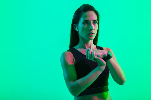 Belle jeune fille de remise en forme portant des écouteurs sans fil isolés sur un mur de néon vert, faisant des exercices d'étirement