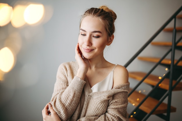 Belle jeune fille rêveuse aux cheveux blonds en robe de soirée en soie beige et cardigan confortable devant les escaliers à la maison