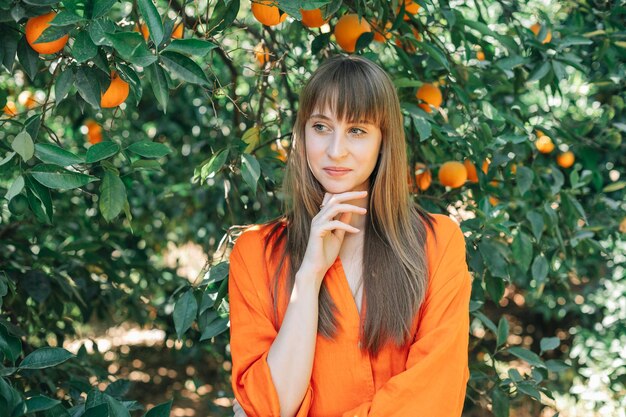 Belle jeune fille en robe orange regarde loin en tenant l'index sous le menton dans le jardin orange