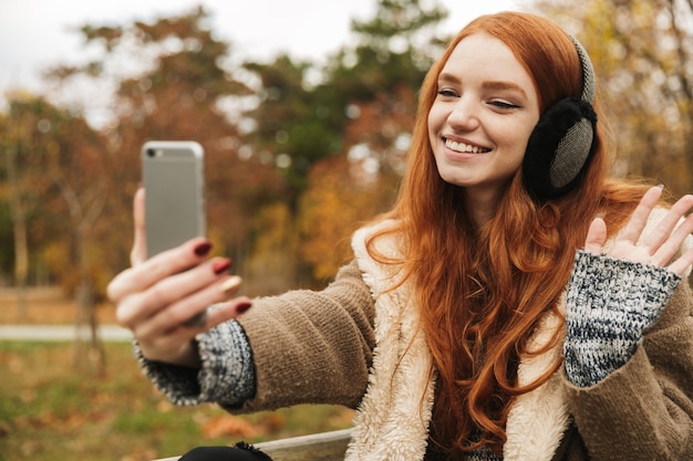 Belle jeune fille rousse écoutant de la musique avec des bandeaux assis sur un banc, prenant un selfie