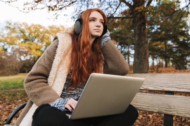 Belle jeune fille rousse écoutant de la musique avec des casques assis sur un banc, à l'aide d'un ordinateur portable