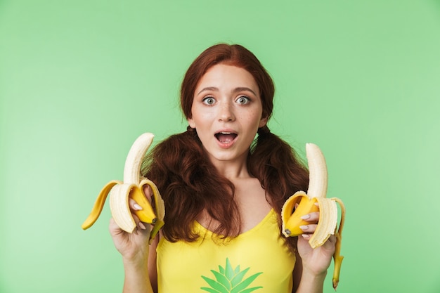 belle jeune fille rousse excitée posant isolée sur fond de mur vert avec des fruits de bananes.