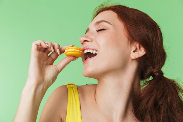 belle jeune fille rousse posant isolée sur fond de mur vert tenant des macarons de bonbons.