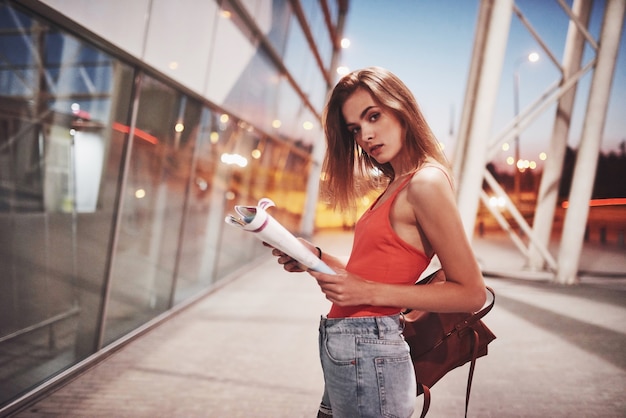 Une belle jeune fille avec un sac à dos derrière ses épaules, debout dans la rue près d'un aéroport. Elle vient d'arriver du reste et très contente!