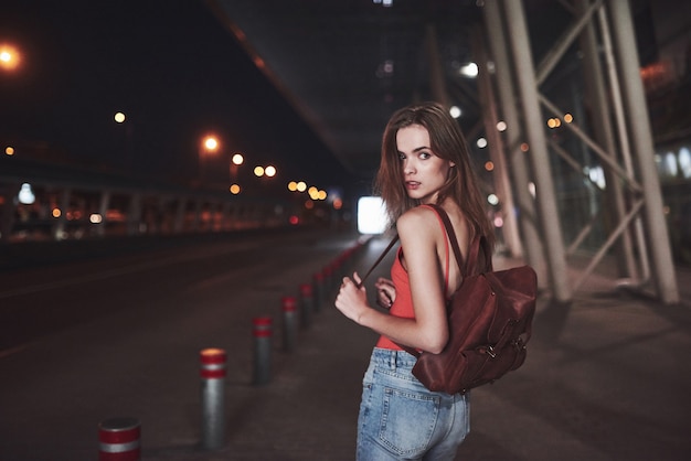 Une belle jeune fille avec un sac à dos derrière ses épaules se tient dans la rue près d'un aéroport ou d'une gare par une chaude soirée d'été. Elle vient d'arriver et attend un taxi ou ses amis.