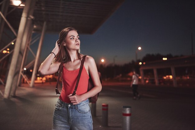 Une belle jeune fille avec un sac à dos derrière ses épaules se tient dans la rue près d'un aéroport ou d'une gare par une chaude soirée d'été. Elle vient d'arriver et attend un taxi ou ses amis.