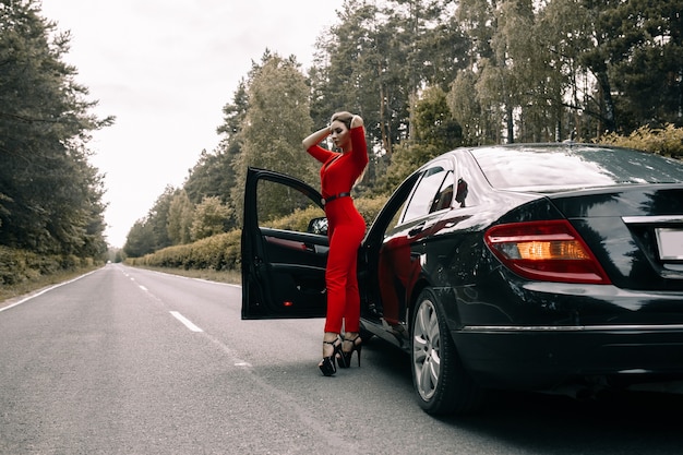Une belle jeune fille en salopette rouge se tient près d'une voiture noire sur une route vide dans la forêt