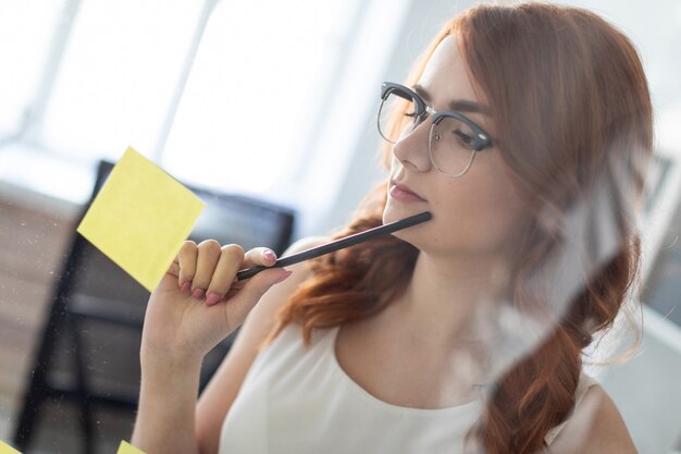 Une belle jeune fille se tient dans le bureau près d'un verre sur lequel des autocollants sont collés