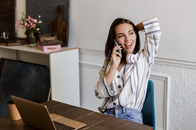 Belle jeune fille souriante assise à la table à la maison, parlant au téléphone mobile