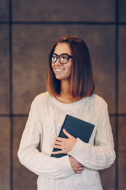 Belle jeune fille souriante dans des verres élégants et un pull blanc avec carnet de croquis d'un mur en béton