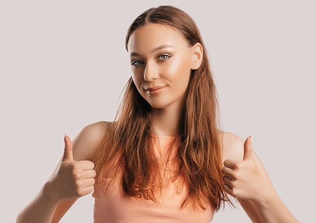 Belle jeune fille souriante et montre le geste du pouce vers le haut avec les deux mains sur un fond blanc isolé. La femme positive indique une idée, un endroit pour la publicité