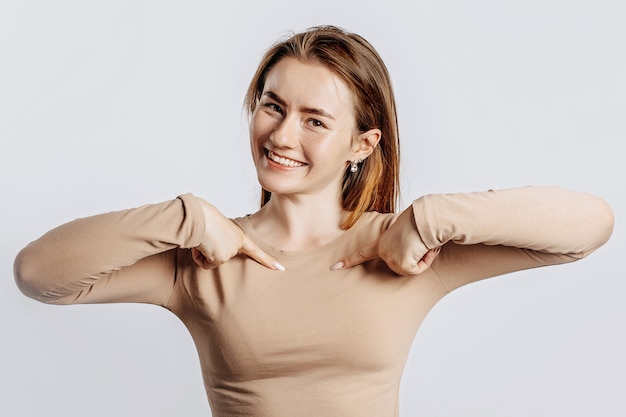 Belle jeune fille souriante et pointant ses doigts sur elle-même sur un fond blanc isolé. Une femme pointe vers une idée, un endroit pour faire de la publicité. Brune positive dans un pull beige.