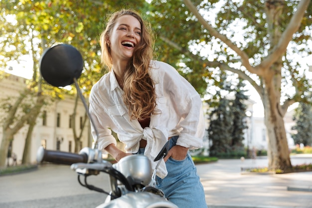 Belle jeune fille souriante portant des vêtements d'été décontractés assis sur un scooter à l'extérieur dans les rues de la ville