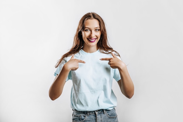 Une belle jeune fille sourit et pointe ses doigts sur elle-même sur un fond gris isolé blanc