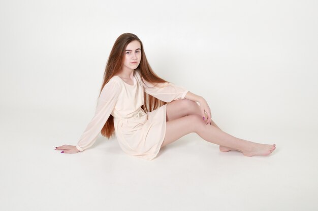 Belle jeune fille tendre vêtue d'une longue robe blanche et aux cheveux longs est assise dans une pose détendue en studio sur fond gris.