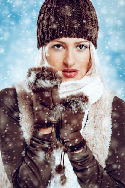 Belle jeune fille en vêtements d'hiver avec un bonnet de laine et des gants de fourrure, colle à l'écharpe en laine dans laquelle se glisse la neige qui tombe.