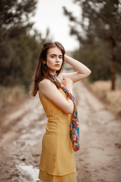 Une belle jeune fille vêtue d'une robe jaune avec une écharpe colorée se dresse sur un chemin de terre au printemps