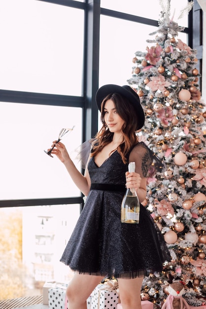 Une belle jeune fille vêtue d'une robe noire et d'un chapeau tient une coupe de champagne dans ses mains