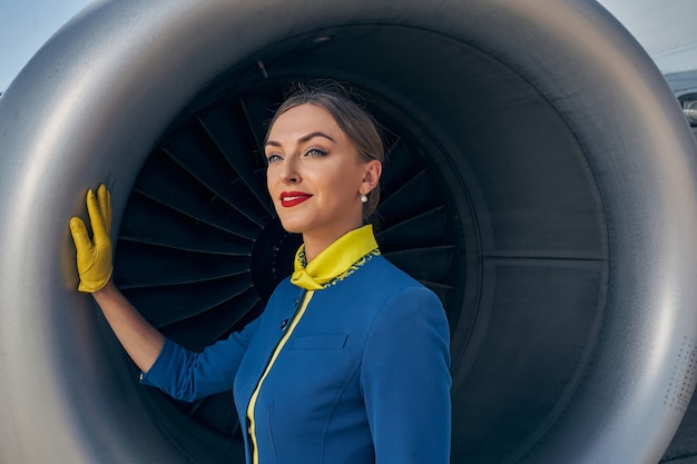 Belle jeune hôtesse de l'air caucasienne rêveuse s'appuyant d'une main sur le moteur d'avion