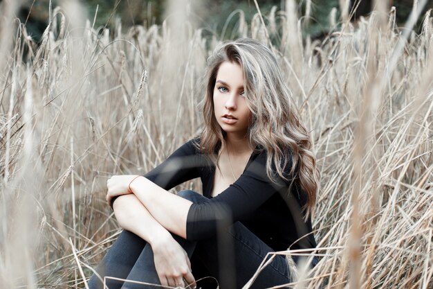 Belle jeune jolie femme blonde en jeans élégants dans une chemise noire à la mode se repose assis dans un champ parmi l'herbe sèche d'automne