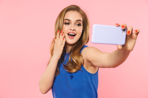 une belle jeune jolie femme excitée posant isolée sur un mur rose prend un selfie par téléphone