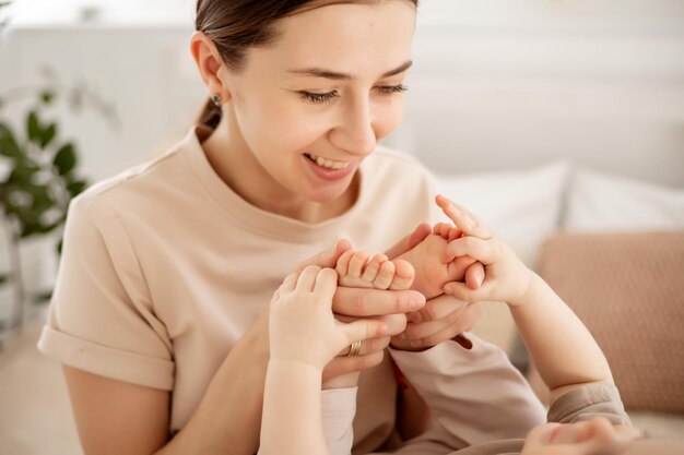 Une belle jeune maman avec un petit garçon étreignant sur le lit à la maison dans la chambre maman tenant les petites jambes du bébé dans ses mains pieds de bébé gros plan