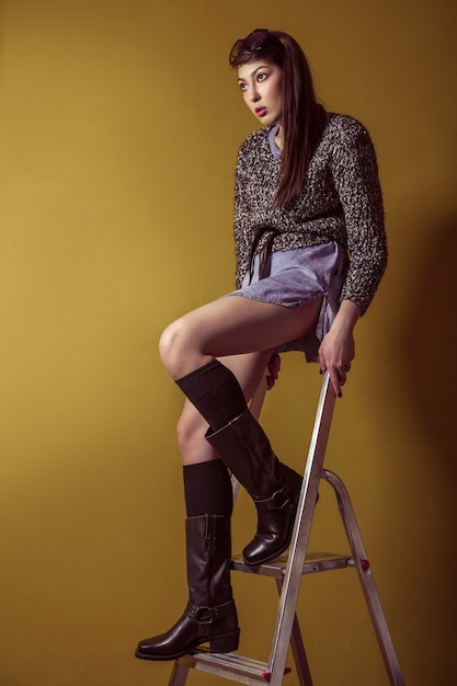 Belle jeune mannequin asiatique en jupe en jean bleu et chemisier noir et maquillage de mode posant assis sur un escabeau et regardant la caméra. Prise de vue en studio. Photo haute couture.