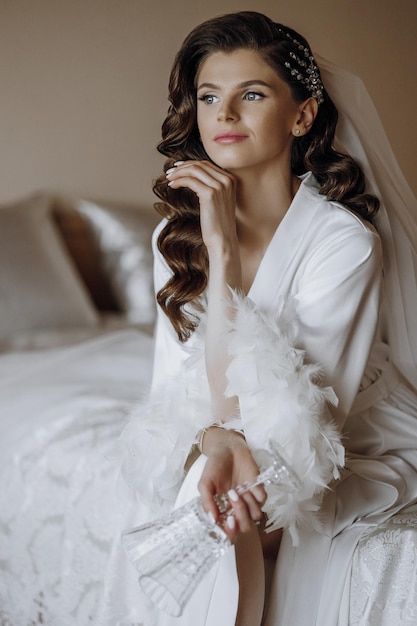 Une belle jeune mariée avec de longs cheveux bouclés dans une robe blanche à la mode est assise sur un lit dans un hôtel et tient des verres dans ses mains une jeune fille belle le matin dans une tenue blanche