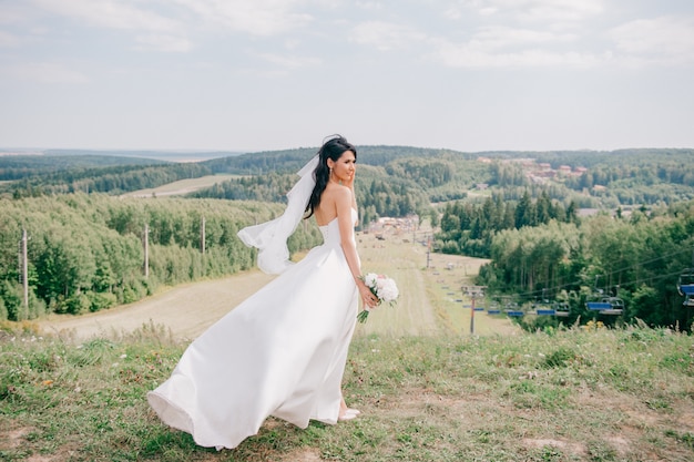 Belle jeune mariée en robe de mariée posant sur la colline à la nature