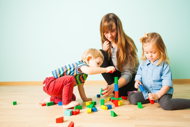 Belle jeune mère aidant ses adorables enfants à construire à partir de blocs de bois colorés. Famille passant du temps à jouer ensemble dans leur maison confortable. Concept parental heureux