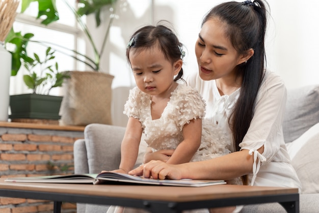 Belle jeune mère asiatique et sa fille assise sur un canapé et lisant une histoire de livre