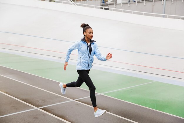 Belle jeune sportive africaine en forme portant des écouteurs sans fil courant au stade