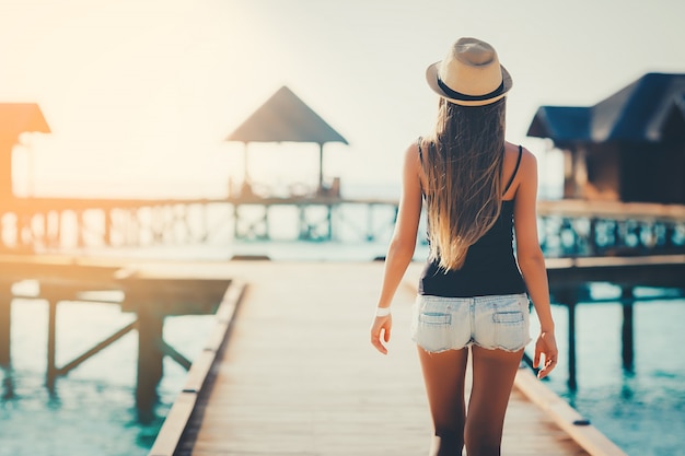 Belle jeune touriste caucasienne avec chapeau marchant sur la jetée dans le lagon au coucher du soleil