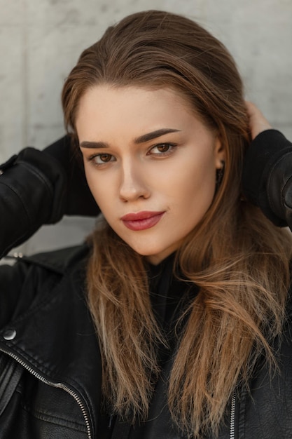 Belle jolie femme modèle avec un visage de beauté frais en veste en cuir noir et sweat à capuche regarde la caméra