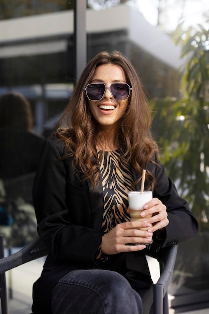 Belle jolie fille à lunettes de soleil est assise à une table dans un café en plein air Une jeune femme d'affaires souriante tient un verre dans ses mains en regardant la caméra
