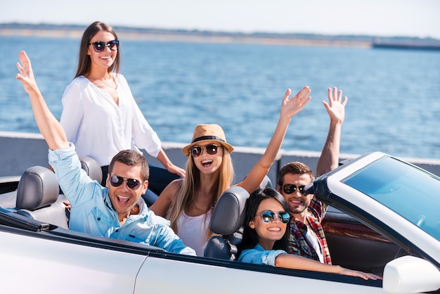 Belle journée pour une balade. Groupe de jeunes heureux profitant d'un voyage en voiture dans leur cabriolet blanc et levant les bras