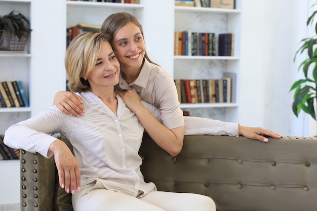 Belle maman d'âge moyen et sa fille adulte s'embrassent et sourient assises sur un canapé à la maison.