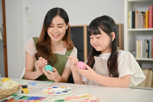 Une belle maman asiatique enseigne et aide sa fille à peindre des aquarelles sur des œufs de Pâques