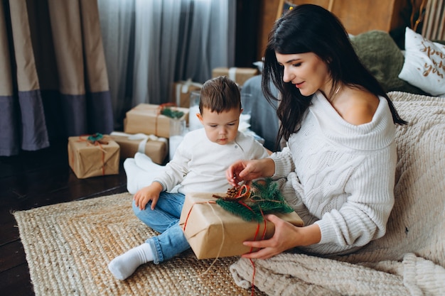 Belle maman aux cheveux bruns et beau garçon fils assis sur le sol et ouvrant des cadeaux dans une boîte sur le