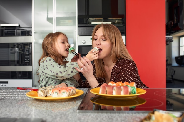 Belle maman caucasienne et sa fille mangeant des petits pains et des sushis lors d'un déjeuner en famille à la maison Livraison de nourriture Cuisine japonaise traditionnelle