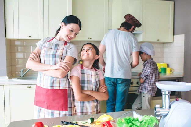 Belle maman et fille se tiennent dos à dos et se regardent. Ils sourient. Les filles se reposent. Les garçons travaillent ensemble derrière. Ils cuisinent.