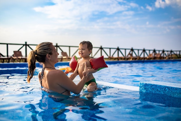 Belle maman joyeuse et joyeuse joue avec son petit fils drôle et mignon avec des manches colorées lumineuses dans une piscine bleu profond avec de l'eau claire et transparente en été