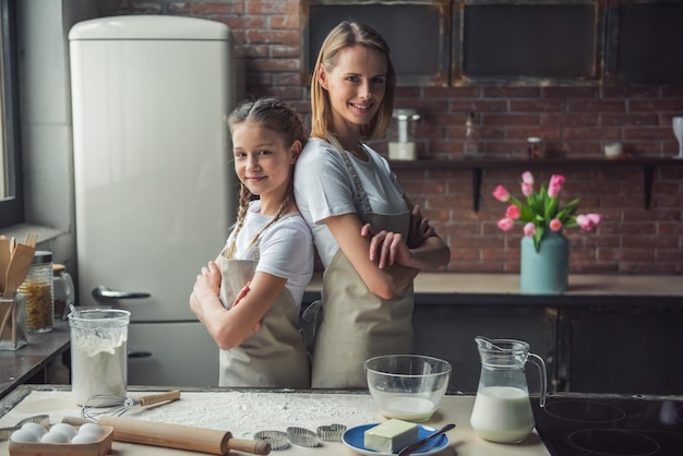 Belle maman et sa fille en tablier regardent la caméra et sourient en se tenant dos à dos dans la cuisine à la maison