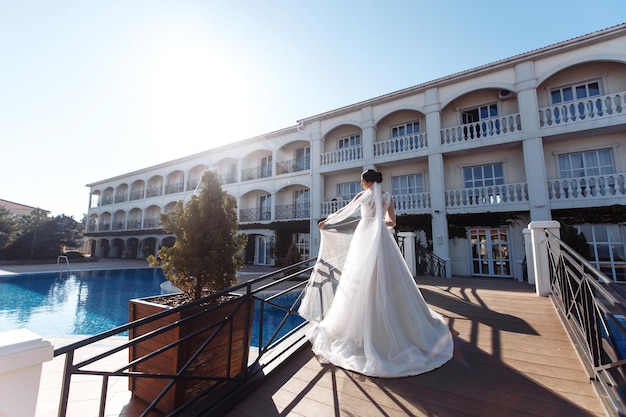 Belle mariée aux cheveux noirs en robe de mariée luxueuse posant sur un balcon avec vue sur la mer