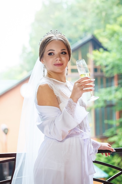 Belle mariée en blouse blanche sur le balcon boit du champagne dans un verre