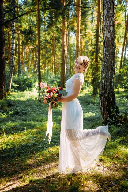 Belle mariée avec un bouquet à la main posant le jour de son mariage parmi les arbres verts