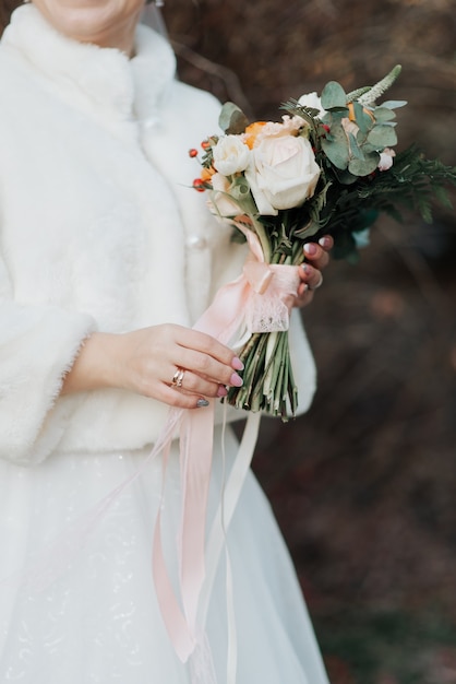 La belle mariée dans un manteau de fourrure blanc tient son bouquet de mariage dans ses mains