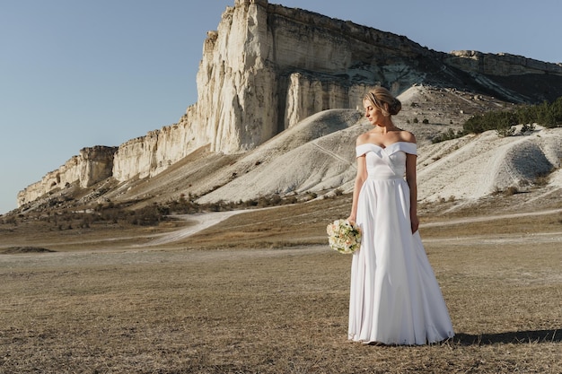 Belle mariée dans une robe de mariée avec un bouquet debout contre un rocher blanc