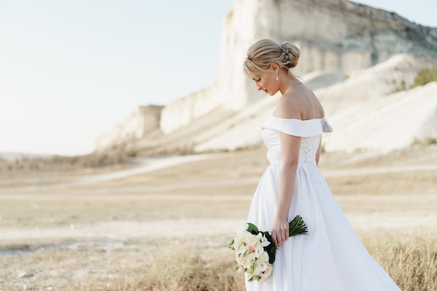 Belle mariée dans une robe de mariée avec un bouquet debout contre un rocher blanc